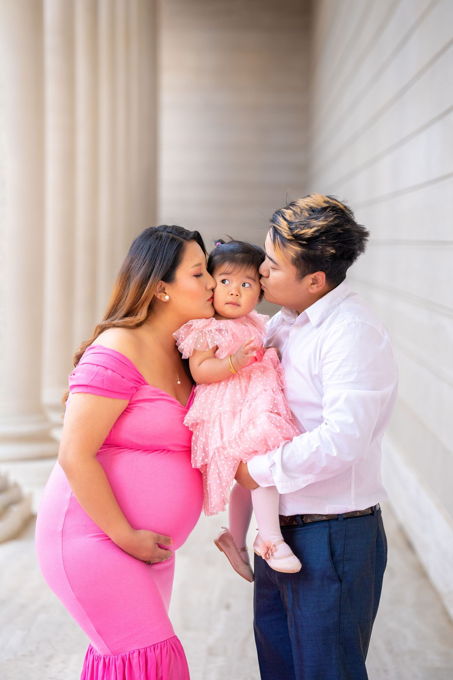 Family maternity photo showing expectant parents and young toddler in pink dress against white columns.