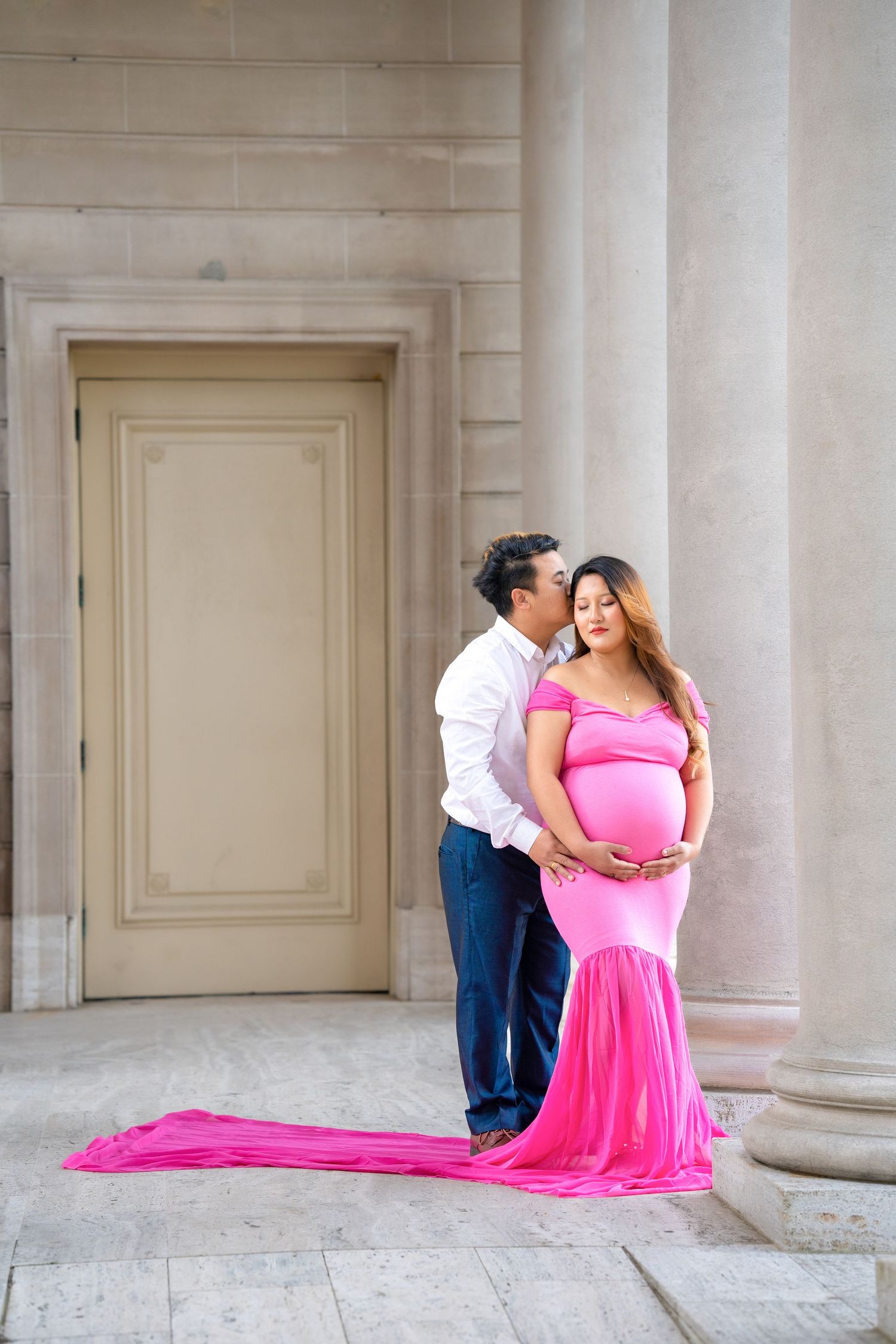 Romantic maternity photo of couple in formal attire posing by classical architecture.