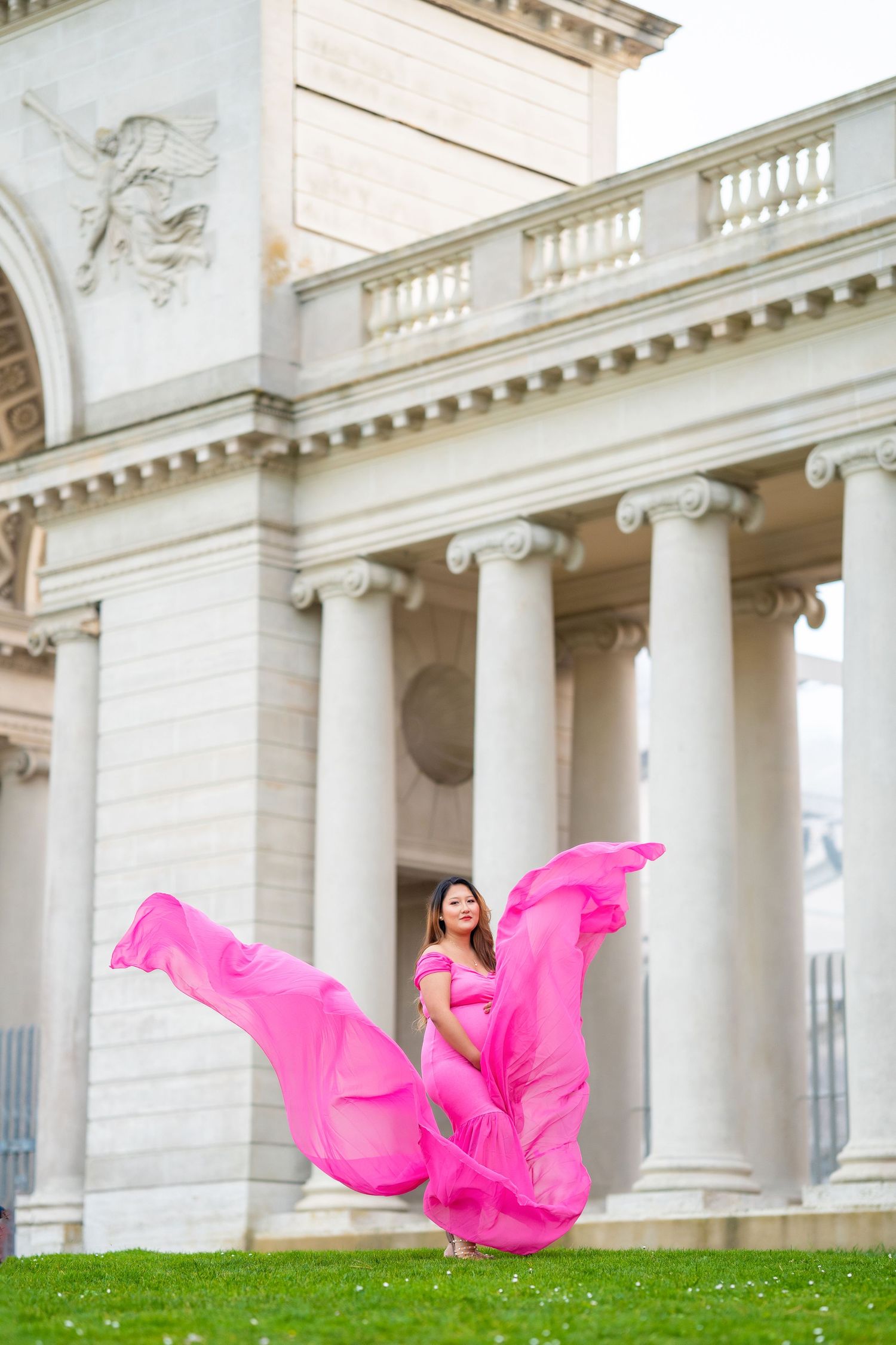 Woman in flowing pink maternity gown twirling on grass in front of columned building.