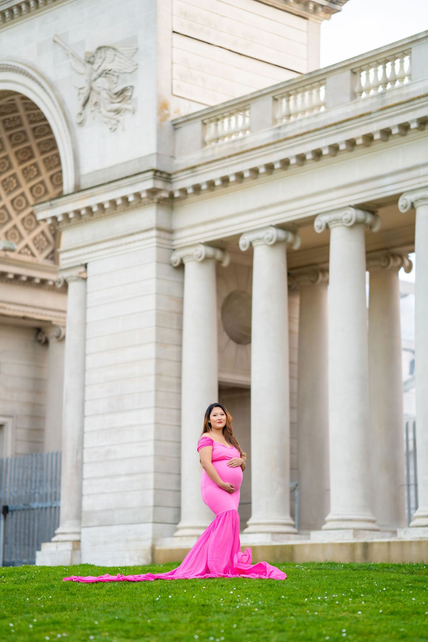 Expectant mother in pink gown poses elegantly against neoclassical architecture with columns.