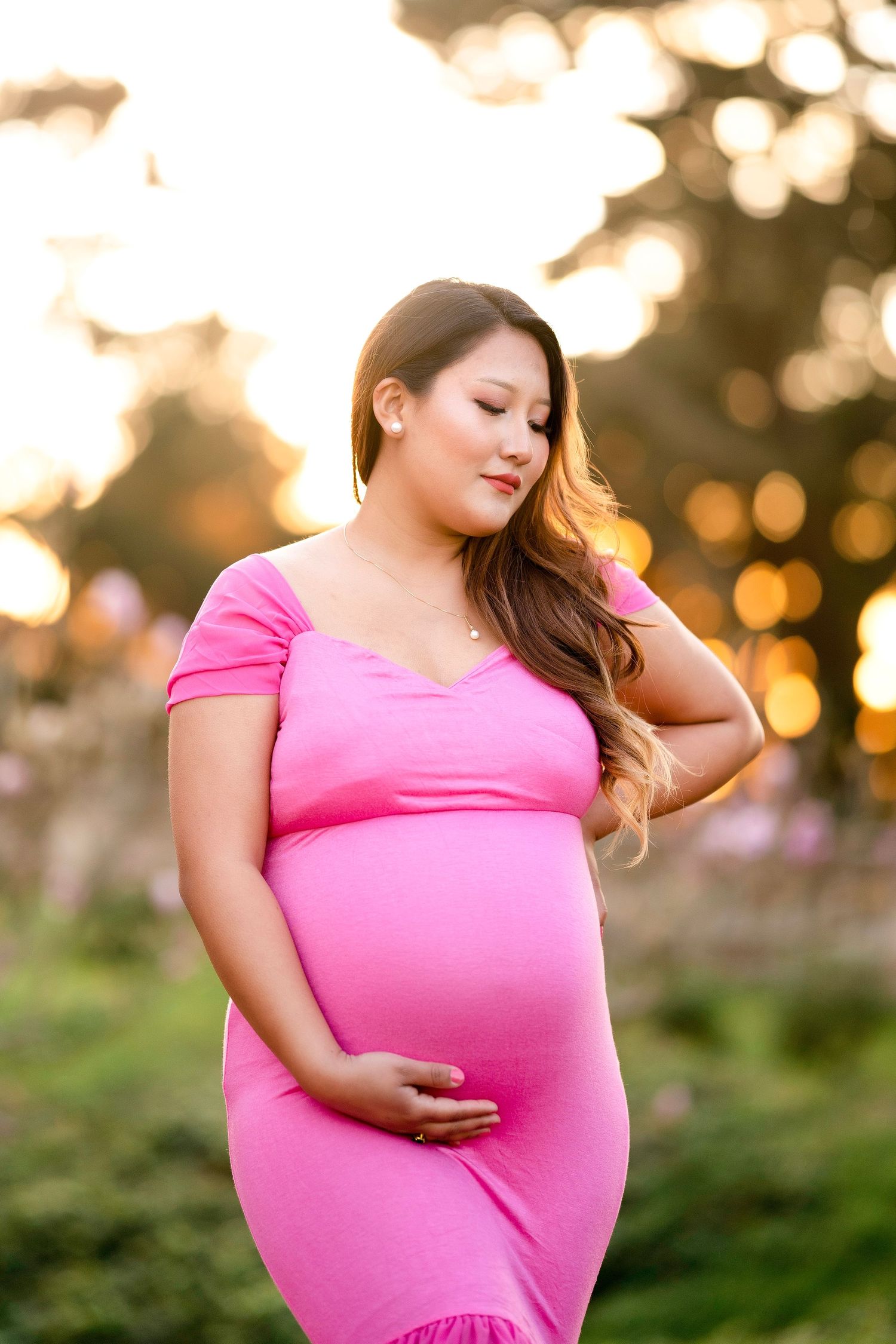 Maternity portrait at sunset featuring woman in fitted pink dress with golden bokeh background.