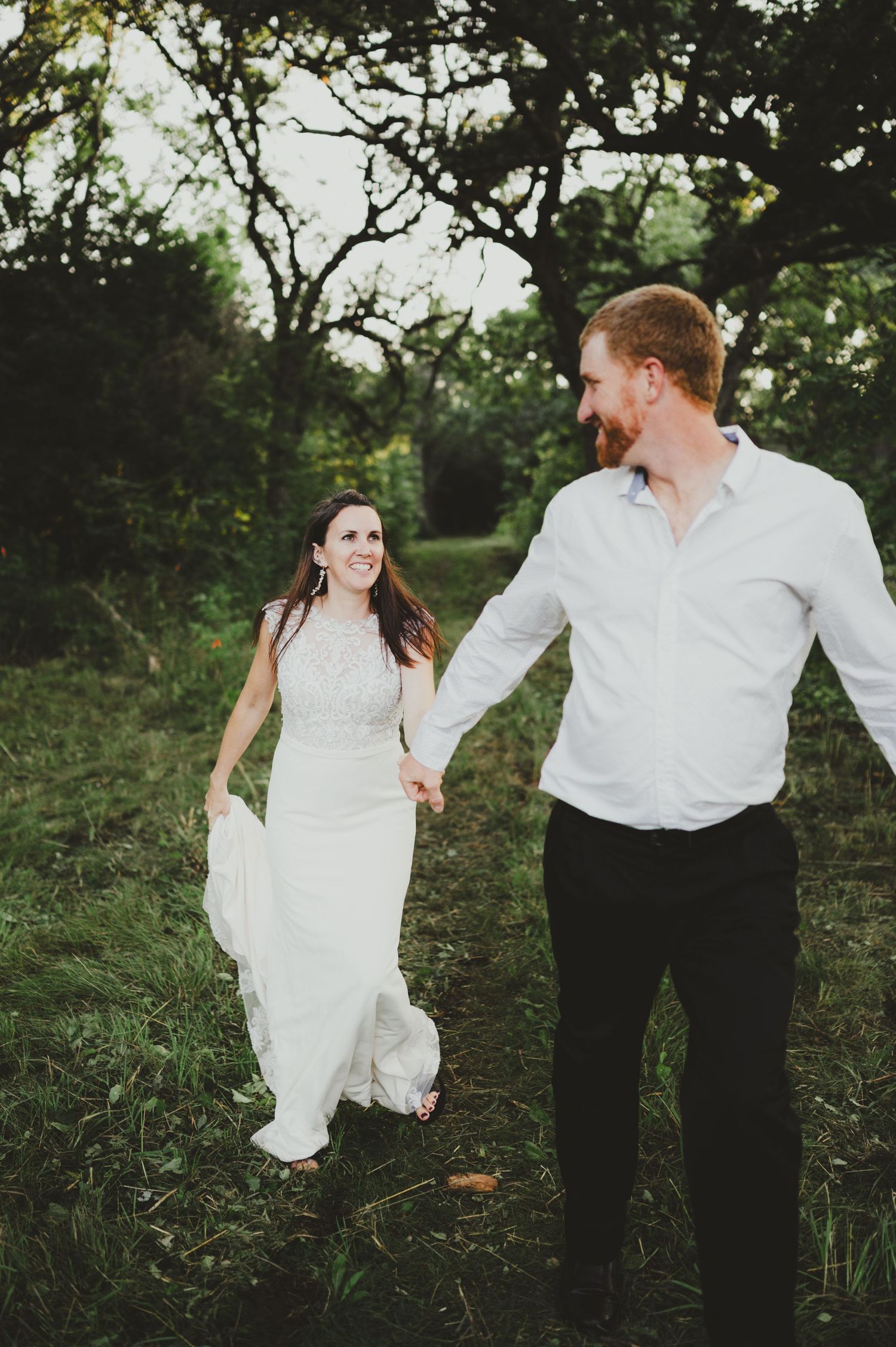 A couple walks hand in hand through a rustic outdoor setting, the bride in a flowing white gown against lush greenery.