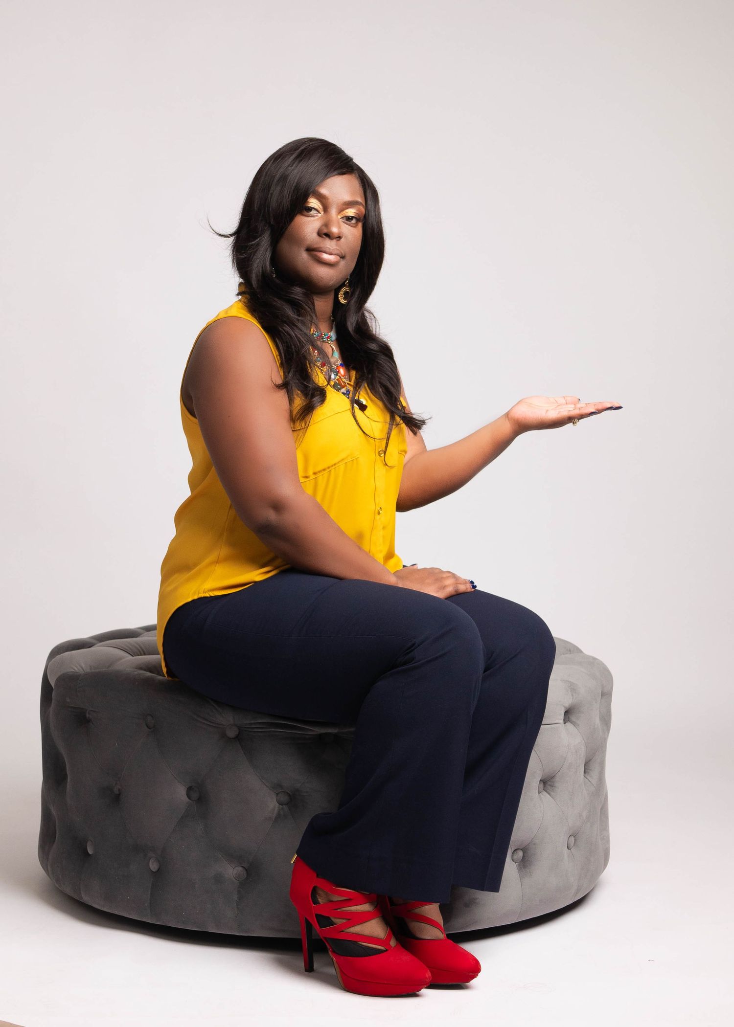 Someone in yellow top sitting on grey ottoman making presentation gesture in studio setting.