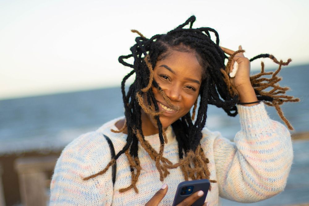 Person with dreadlocks smiling while playing with hair outdoors.
