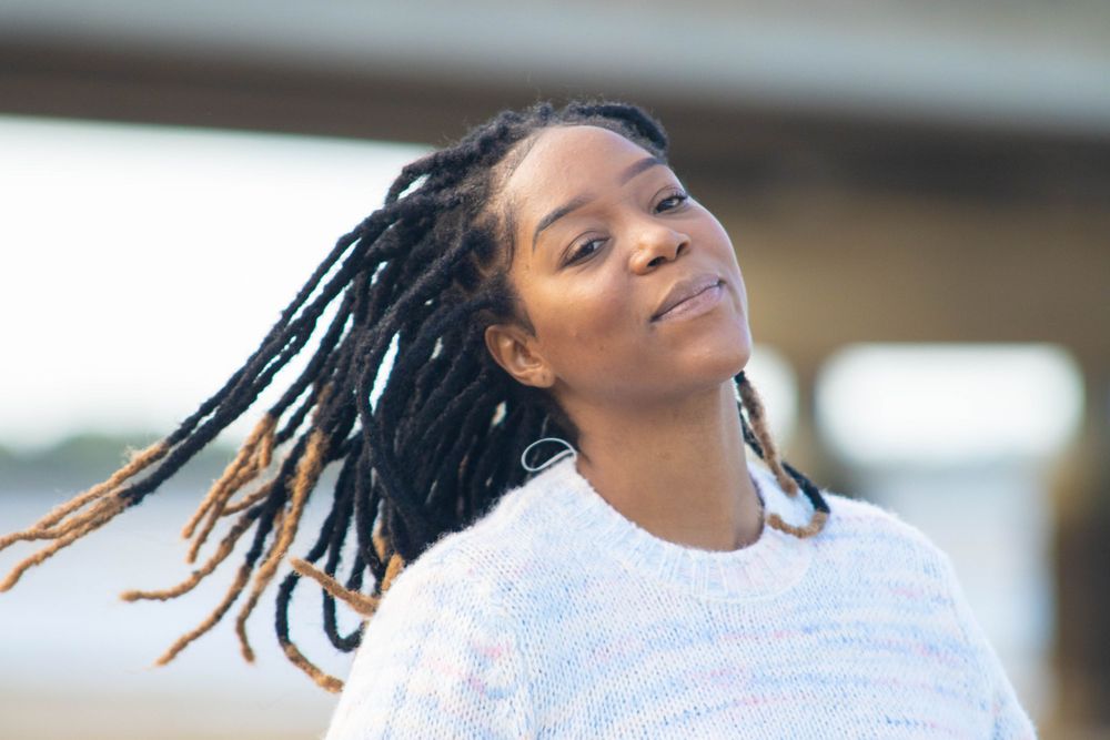 Person with dreadlocks wearing white sweater outdoors smiling.