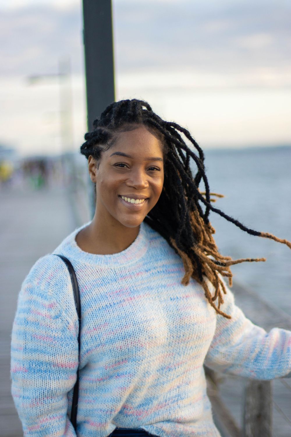 Person in cozy white sweater smiles while standing on a boardwalk by the water.