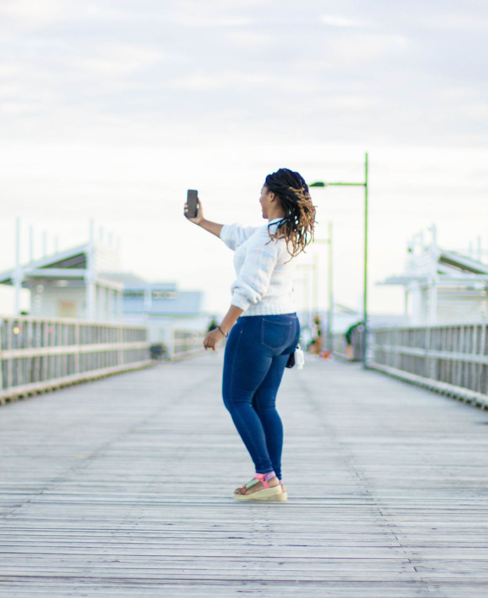 Person taking a selfie while standing on wooden pier with water and buildings in background.