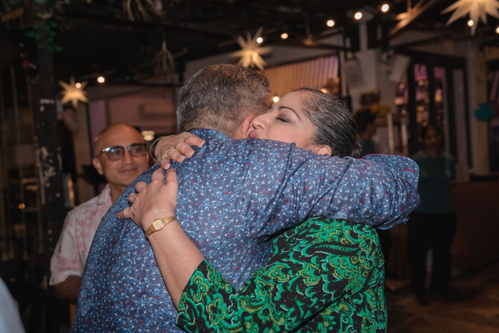 Two people share a warm embrace at a festive evening gathering with string lights in the background.