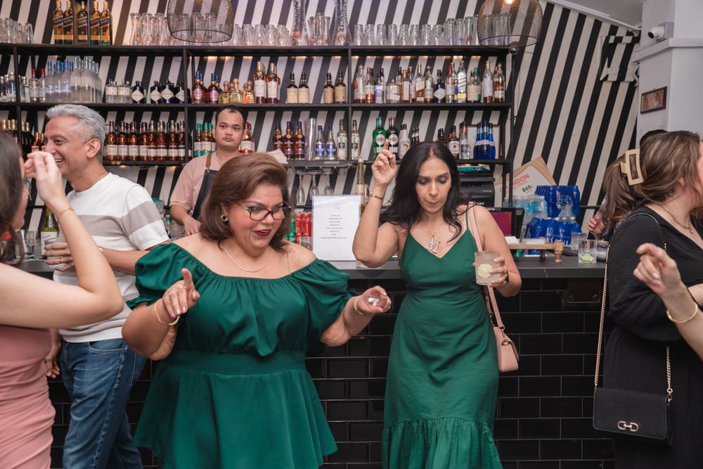 Group of partygoers in green dresses dancing and celebrating at a bar with bottles displayed in background.