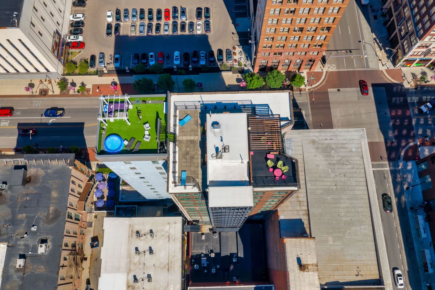Aerial view of urban rooftops showing parking lots, a small green space, and building shadows in a downtown setting.