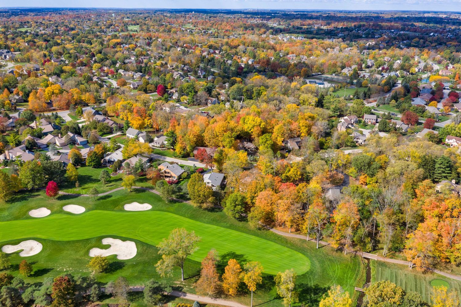 Aerial drone view of a golf course surrounded by colorful fall foliage and suburban homes in a Muirfield neighborhood.