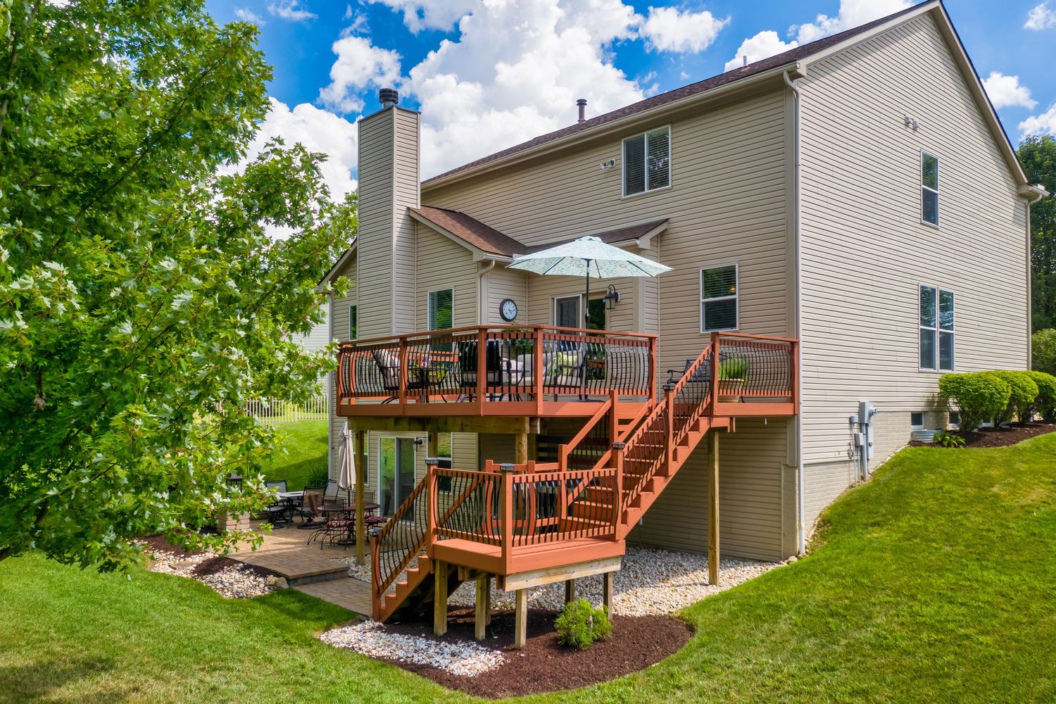 Elevated outdoor deck with wooden stairs leads down to patio space behind beige two-story house with green landscaping.