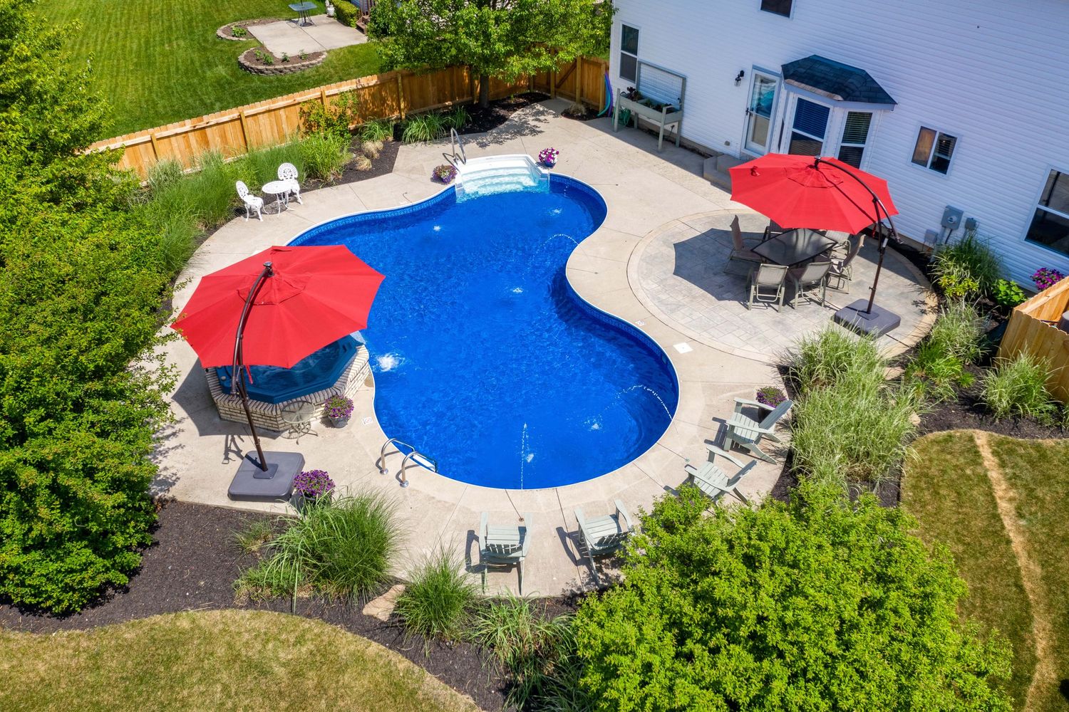 Aerial view of a curved blue swimming pool with red umbrellas and patio furniture surrounded by green landscaping.