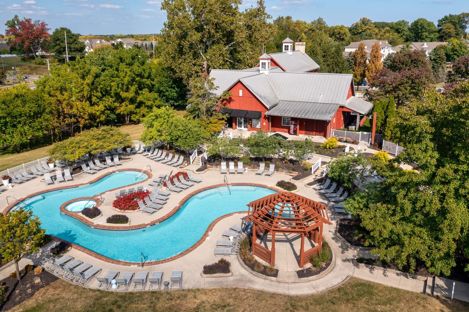 Aerial view of a curved swimming pool with lounge chairs and a gazebo at a multi-family apartment property surrounded by trees.