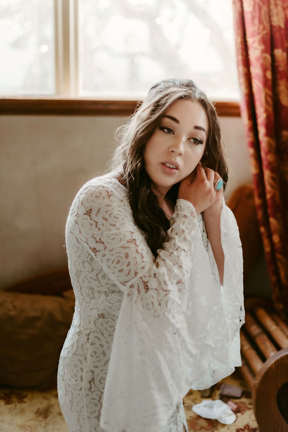 Woman in white lace dress poses near window in soft natural light for bridal portrait.