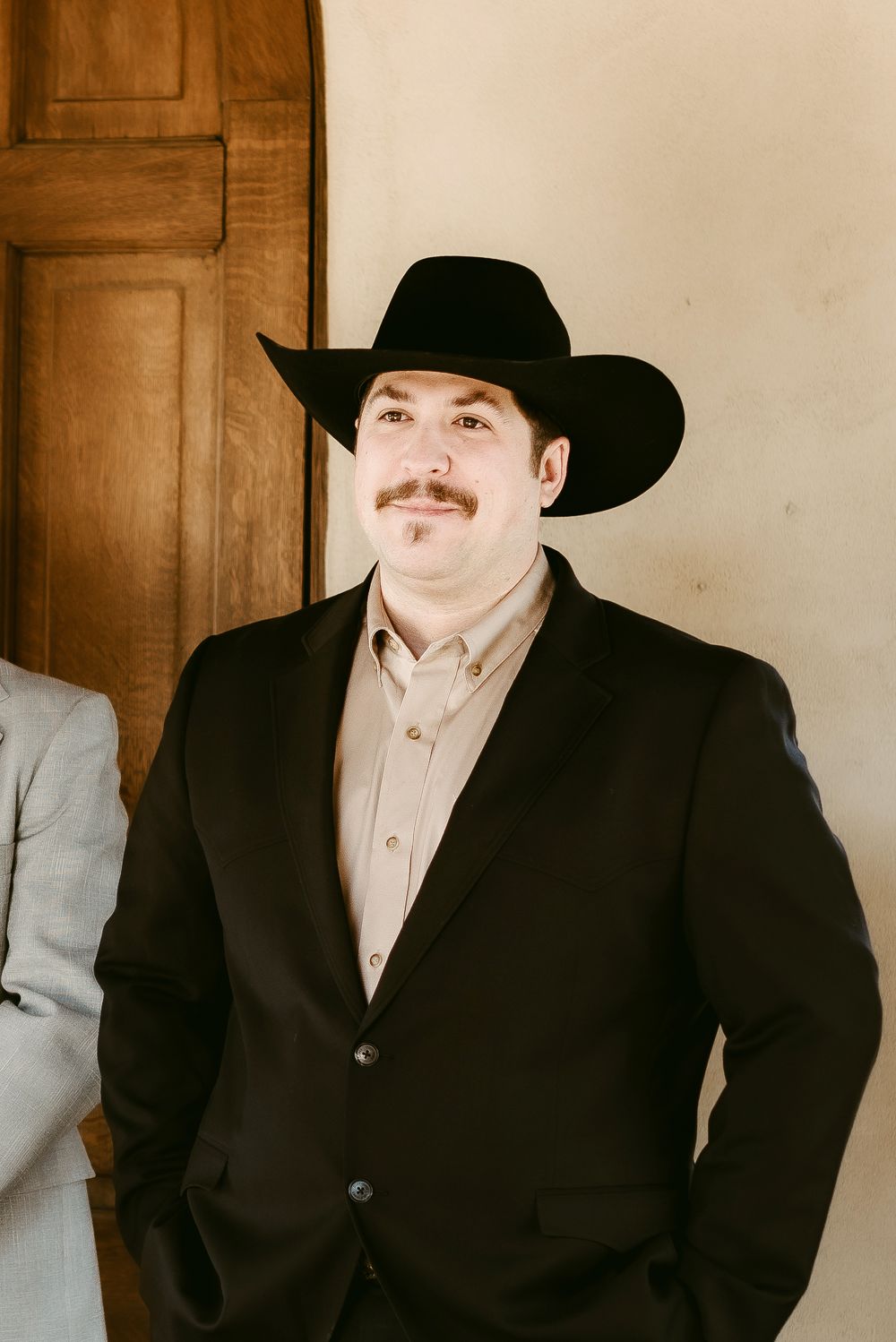 Person in black cowboy hat and suit jacket poses for portrait against wooden door.