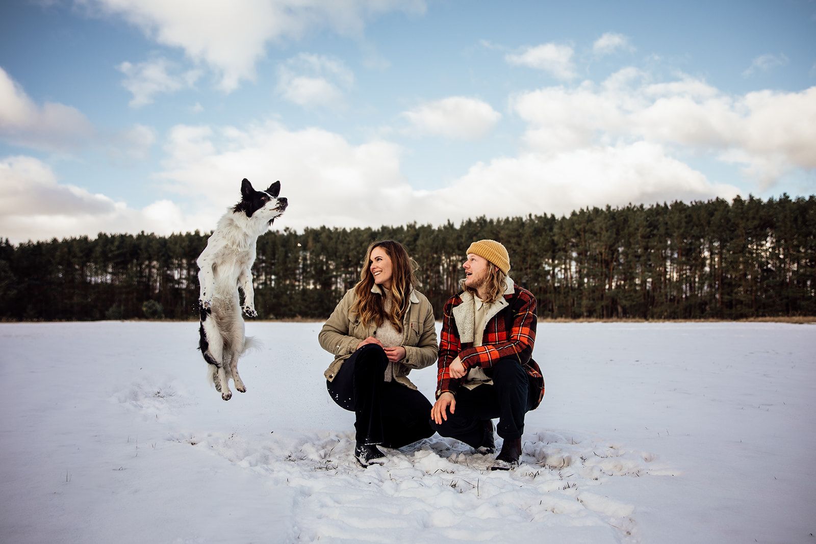 A black and white dog leaps joyfully in the snowy field while two people crouch nearby in winter coats. Dog Photography Berlin