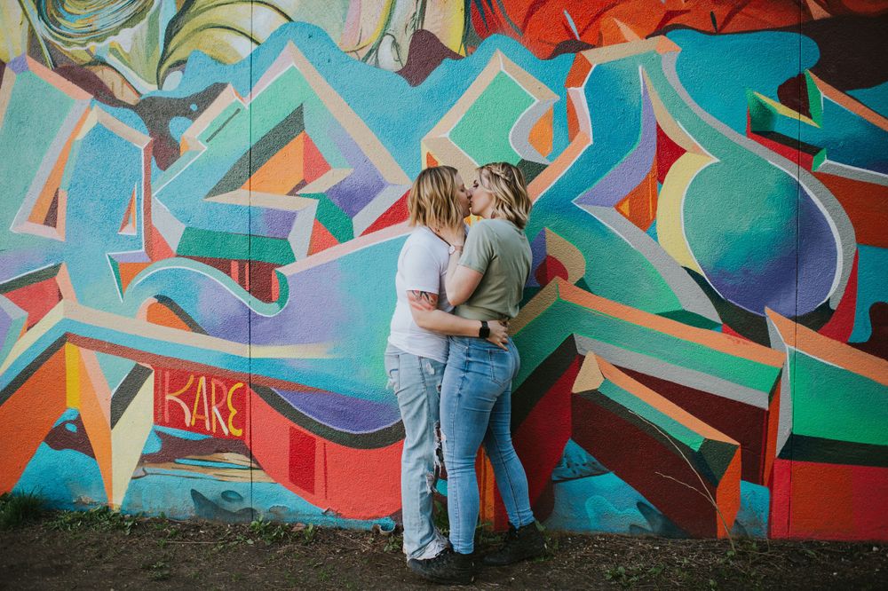 Two women embrace in a passionate kiss in front of a graffiti wall.