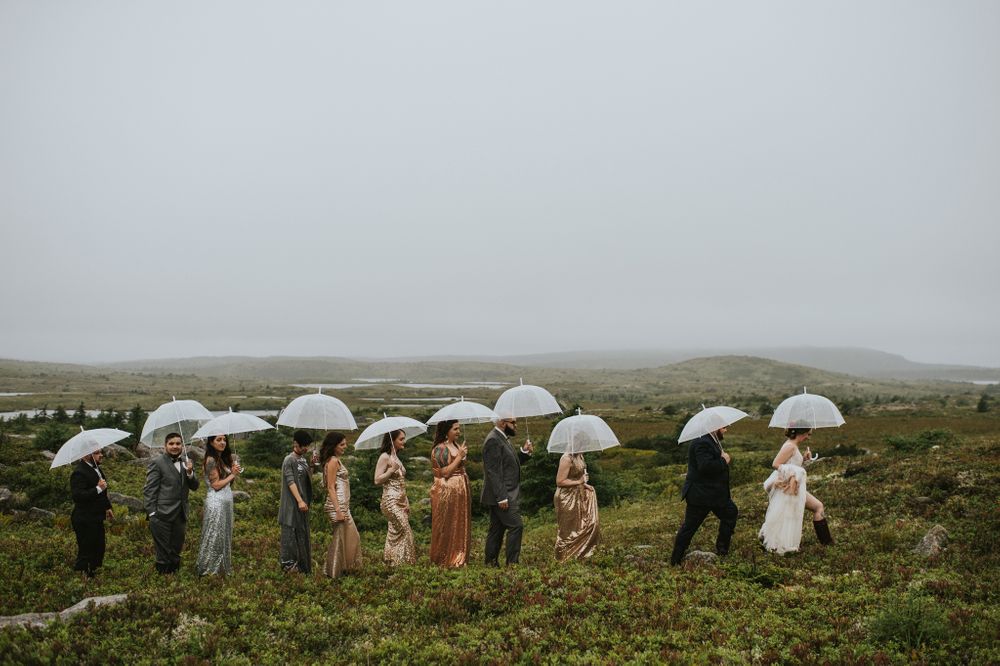 A brightly dressed bridal party walk across a barren marsh in a straight line holding umbrellas.