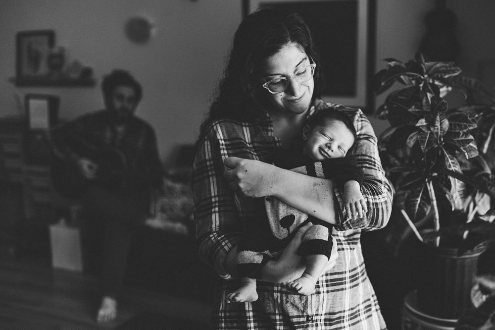 A mother holds her newborn child as the father plays guitar in the background.
