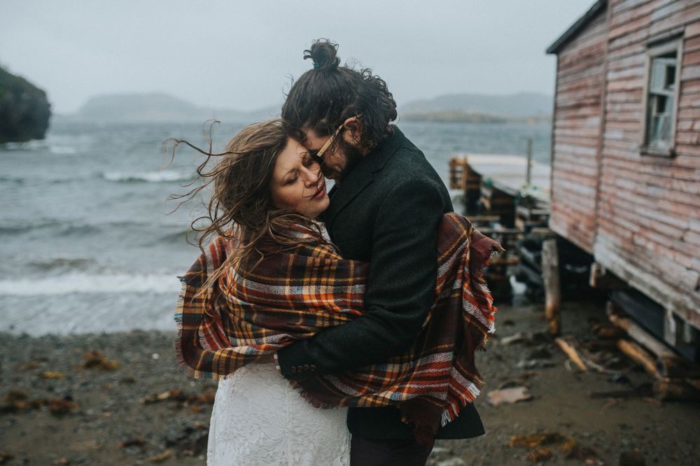 Bride wrapped in plaid and groom embrace on windy Newfoundland shore.