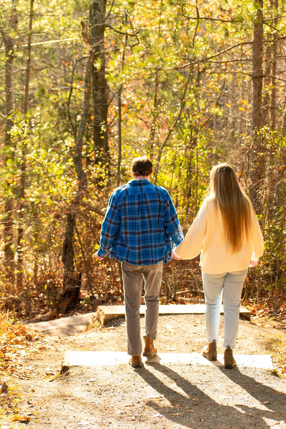 A couple walking hand in hand down a sunlit forest path during autumn.