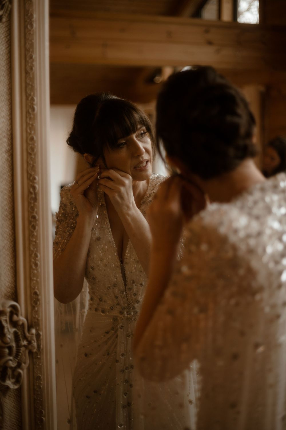 A bride in a sequined wedding dress puts on earrings while looking in an ornate mirror.