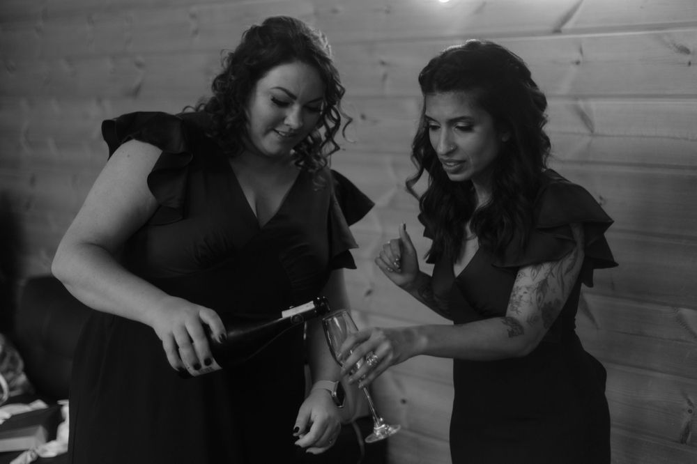 Two bridesmaids in black flutter-sleeve dresses share a moment during wedding preparations.