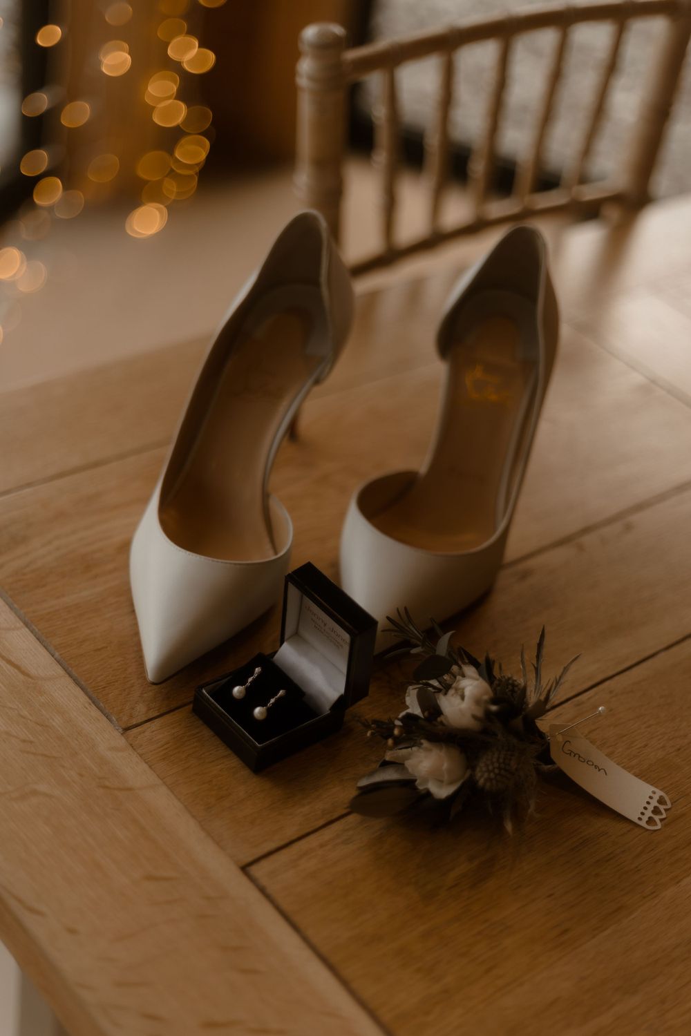 White bridal heels with wedding rings in black box and floral decor on wooden floor.