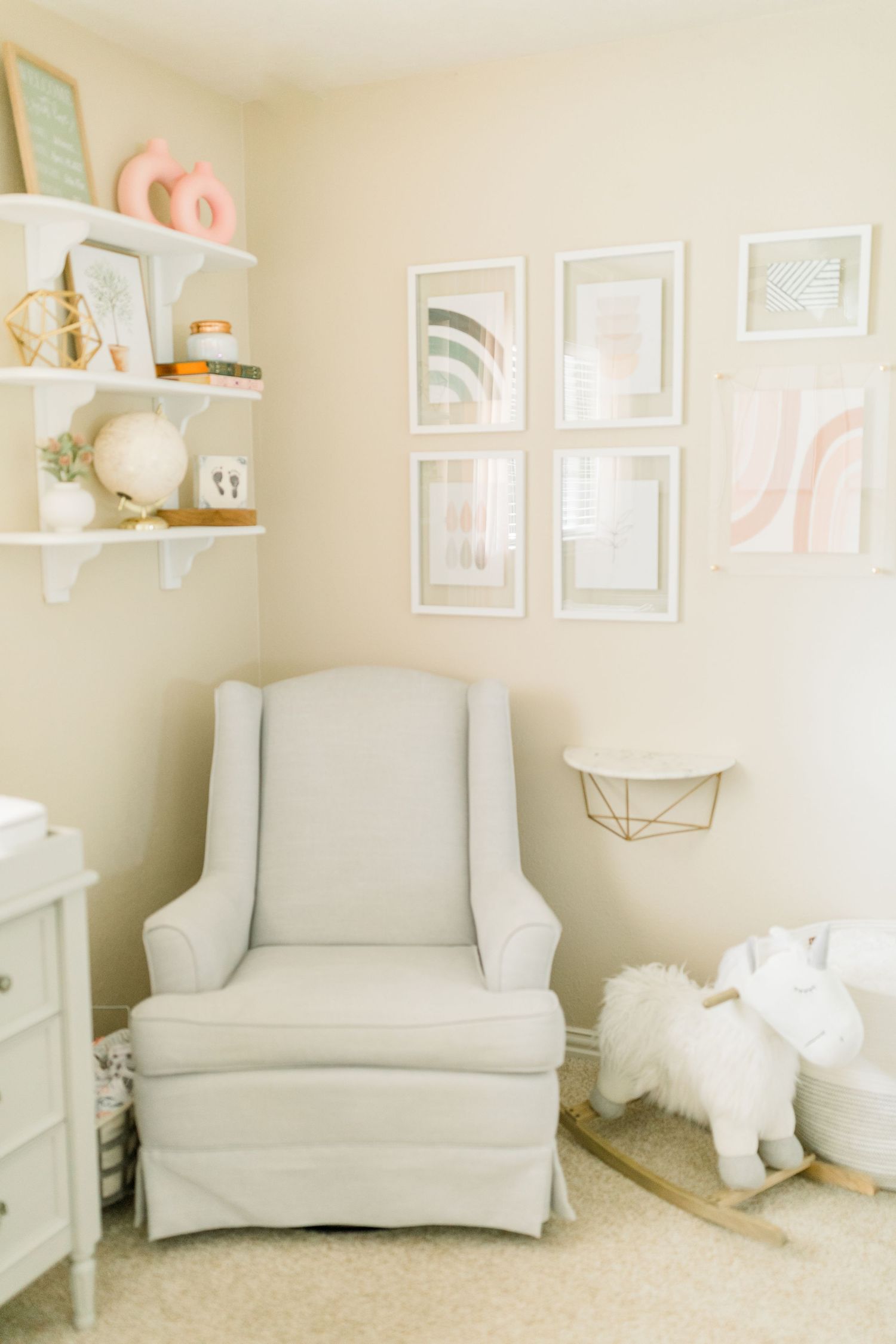 Soft neutral nursery corner with white armchair, floating shelves, and pastel wall art.