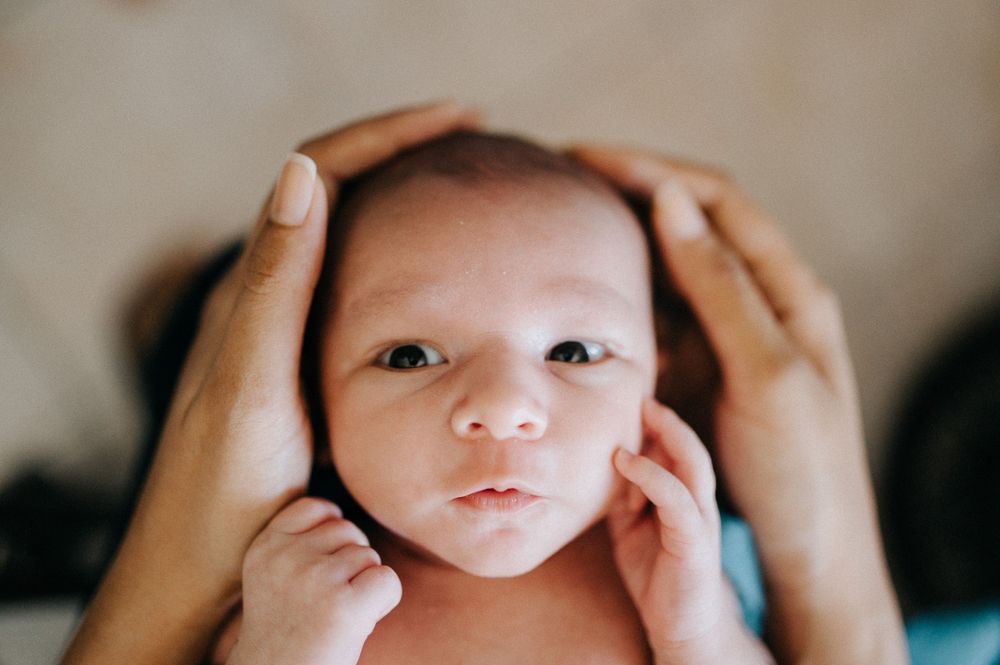 A close-up portrait of a tiny infant's face framed by caring hands from above.