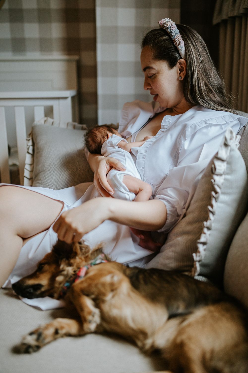 Parent nursing newborn on couch while dog rests beside them.