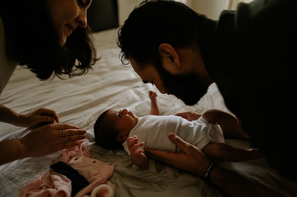 Parents leaning over infant on bed in warm, intimate indoor setting.