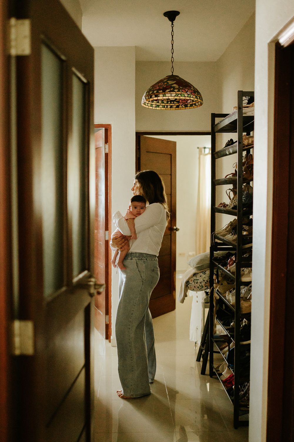 Parent holding baby in hallway with stained glass pendant light and shoe storage.