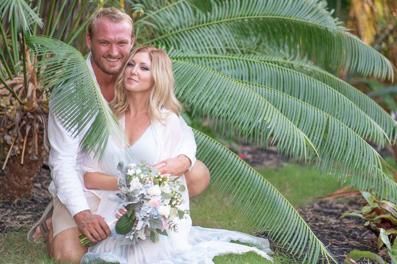 Newlyweds embrace near tropical palm fronds during their beach wedding photoshoot.