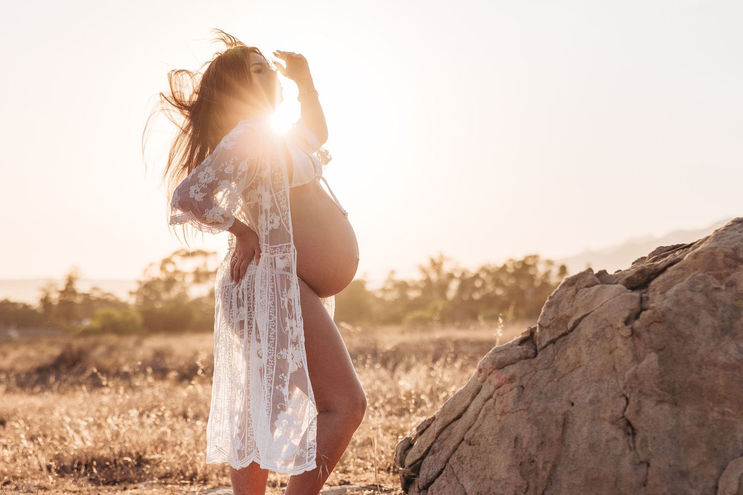 Dreamy silhouette maternity photo with sunburst and flowing white lace dress in golden field.