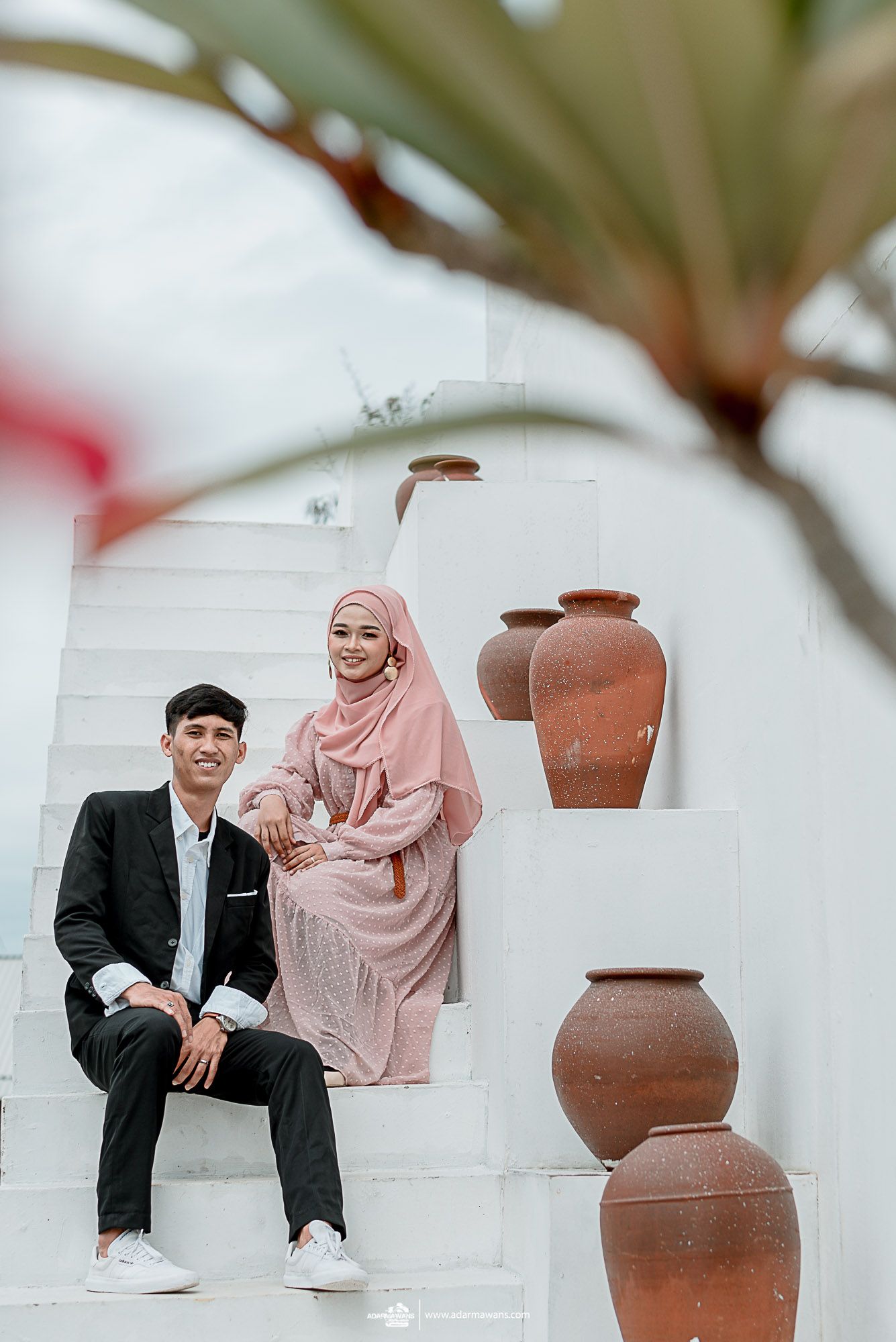 A couple in formal attire poses on white steps with decorative terracotta pots.