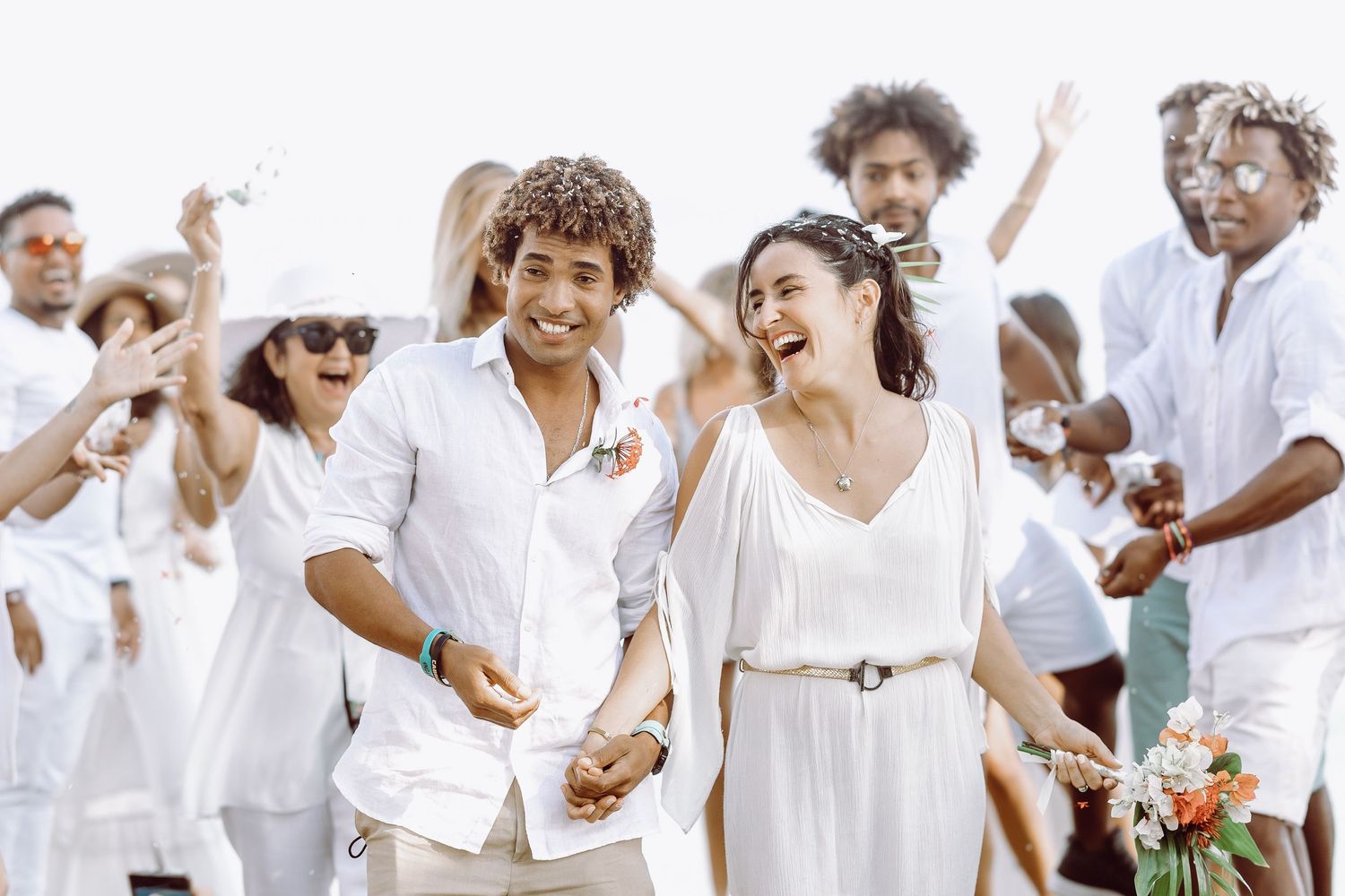 Wedding party guests in all white outfits celebrate joyfully on the beach.