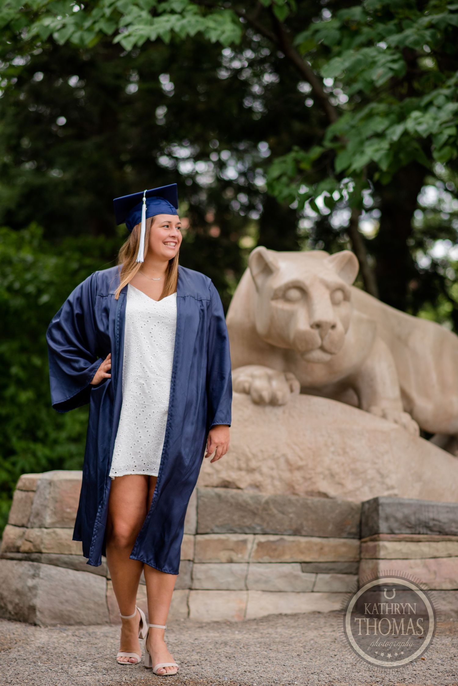Graduate in navy blue gown strikes confident pose beside stone lion statue.
