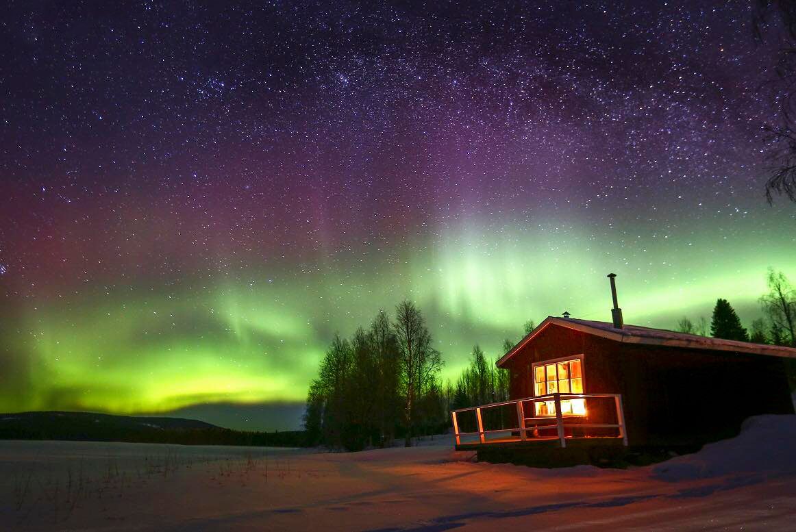 Vibrant green and purple aurora borealis illuminating the night sky above a cozy lit cabin in snowy wilderness.