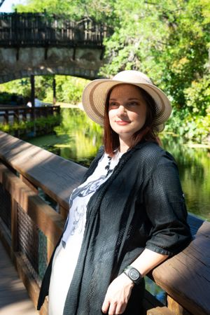 Person in black jacket and sun hat relaxing on wooden bench by serene pond surrounded by lush greenery.