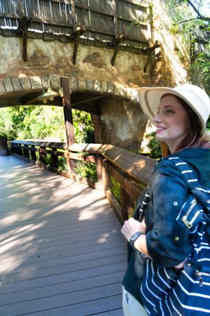 Person wearing striped shirt and hat exploring covered wooden walkway in dappled sunlight.