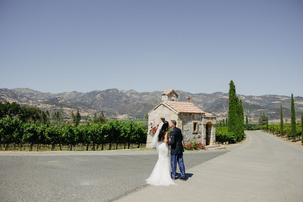 A couple walks along a scenic vineyard path leading to a small chapel with mountains in the background.