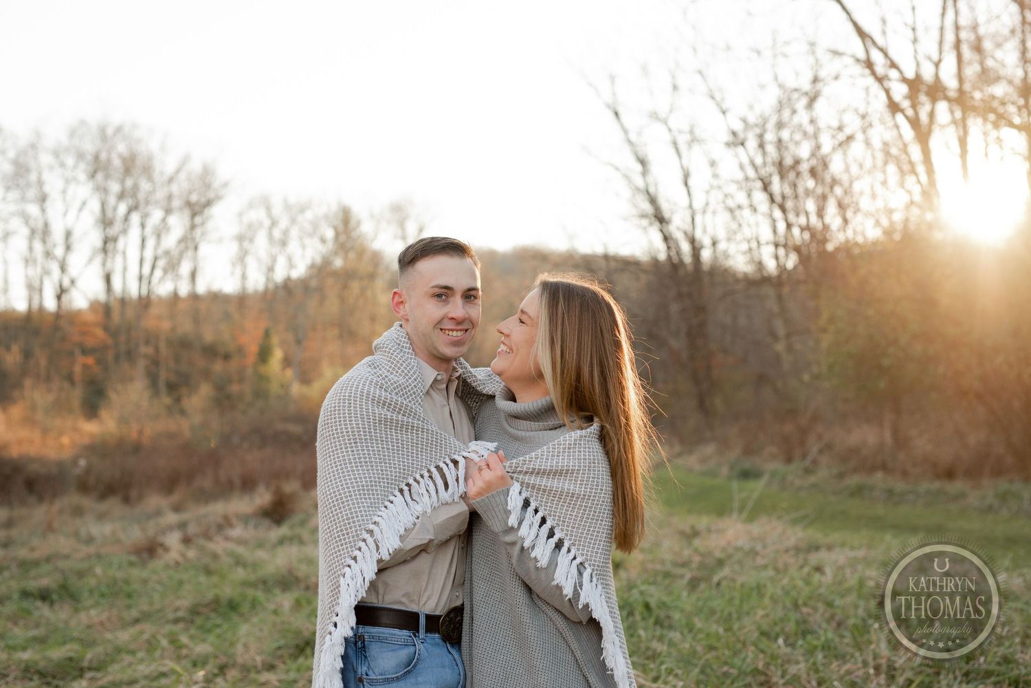 A couple wrapped in a blanket shares a loving moment during sunset in a fall field setting.