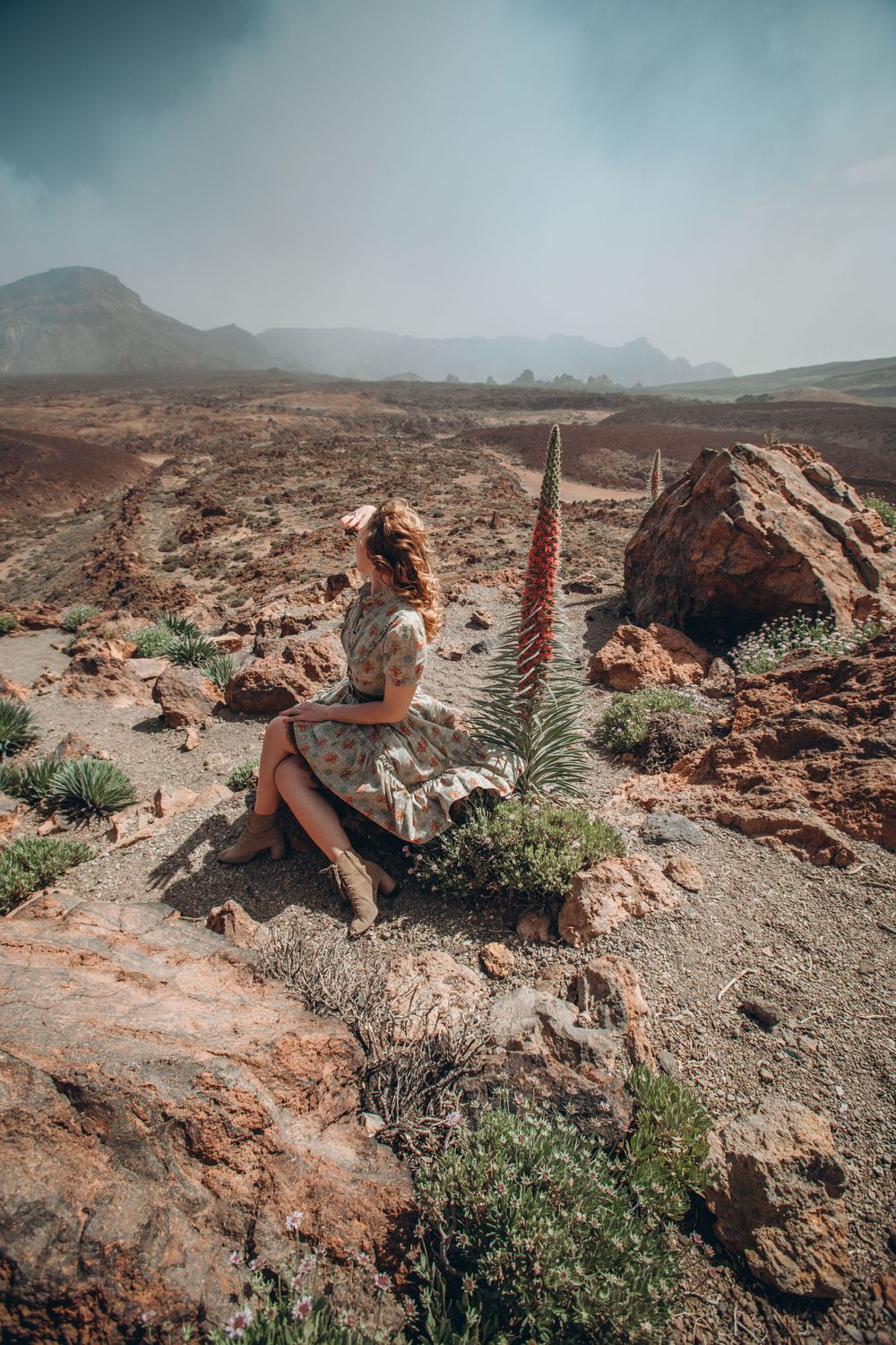 Person relaxing on rocky desert terrain with mountains and desert plants in the background.