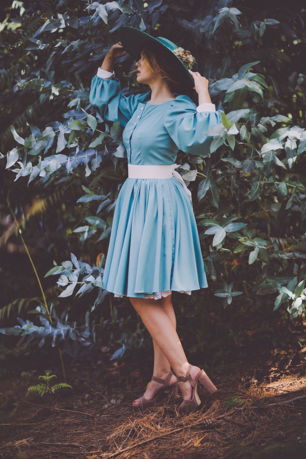 Someone in a blue vintage-style dress posing playfully among garden foliage.