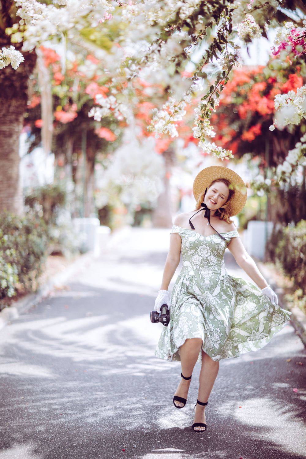 A flowing floral dress twirls beneath blooming cherry blossoms on a sunny pathway.
