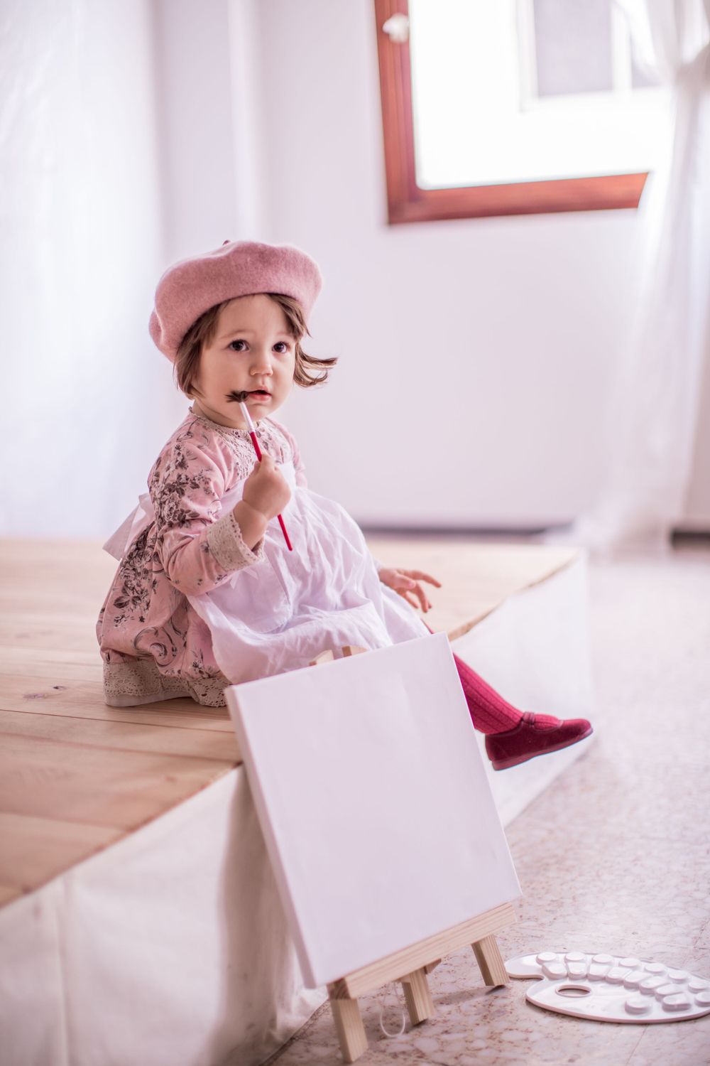 Small child in pink dress and beret plays with white box on wooden floor in bright sunny room.