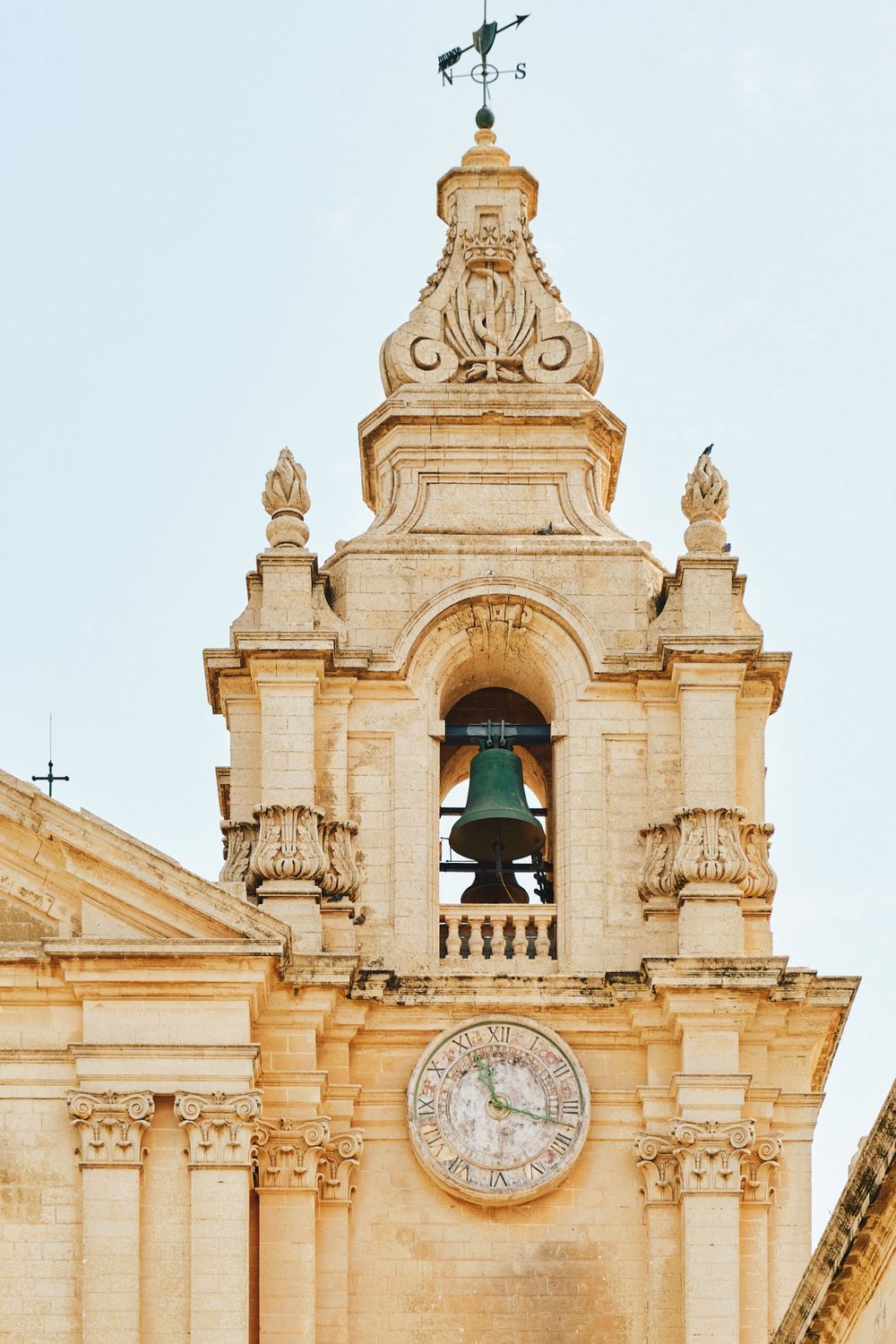 Bell tower with ornate clock face and baroque architectural details against blue sky.