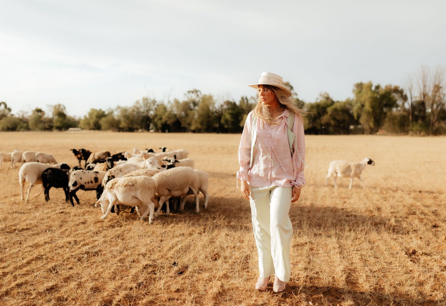 A woman in a stylish pastel outfit and wide-brim hat walks through a sunlit paddock, surrounded by a flock of sheep.